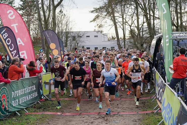 Startszene eines Wettlaufs mit vielen L&auml;ufern und Zuschauern, umgeben von Werbebannern und B&auml;umen.