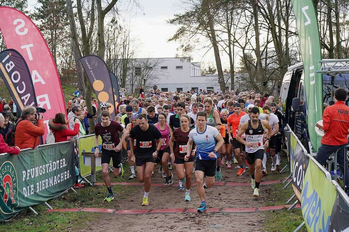 Startszene eines Wettlaufs mit vielen Läufern und Zuschauern, umgeben von Werbebannern und Bäumen.