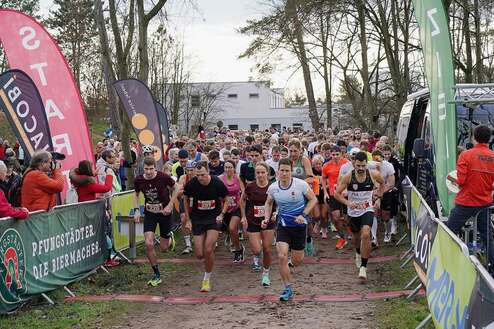 Startszene eines Wettlaufs mit vielen Läufern und Zuschauern, umgeben von Werbebannern und Bäumen.