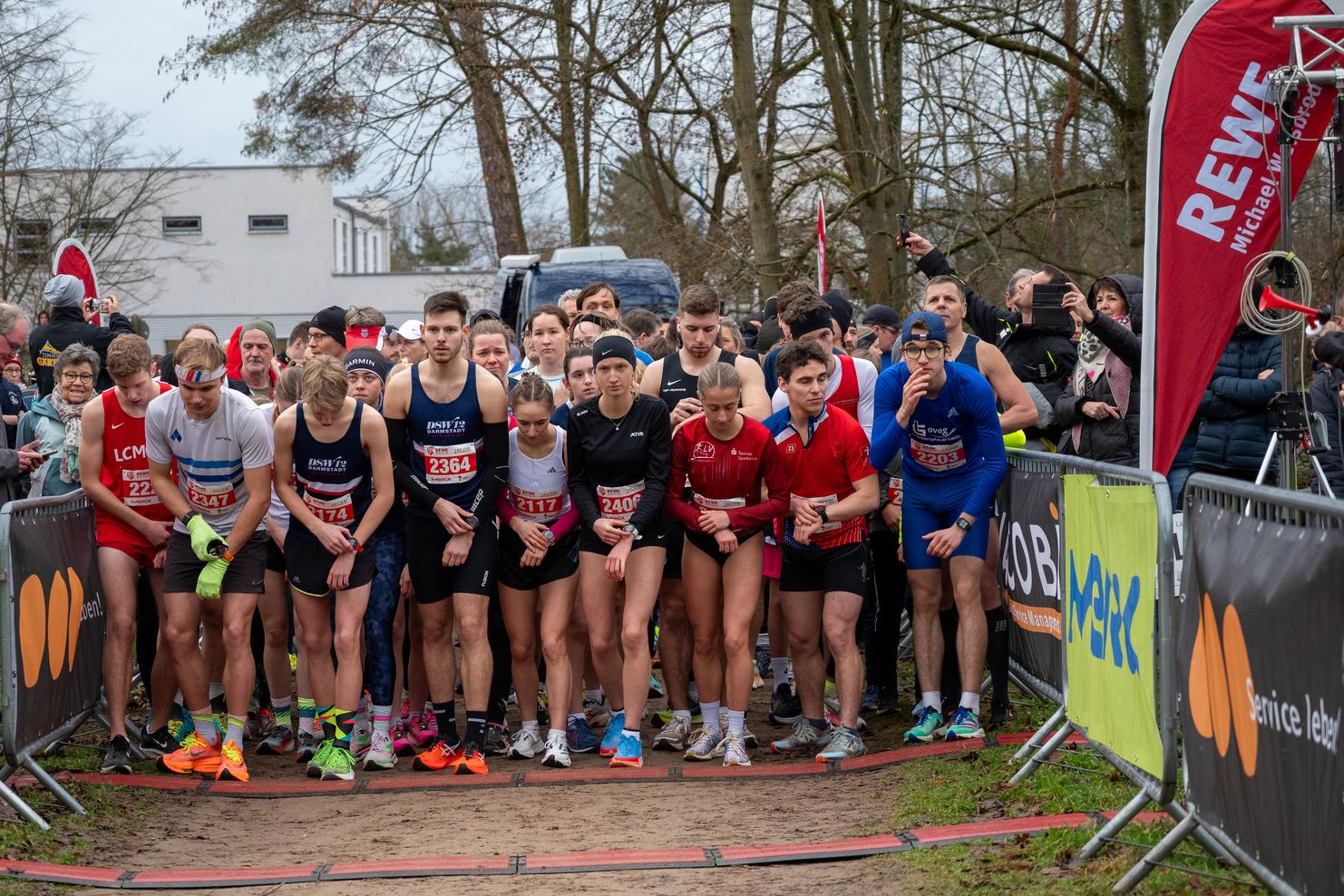 Gruppierung von Läufern am Start eines Wettlaufs, umgeben von Zuschauern, mit Markierungen und Banner im Hintergrund.