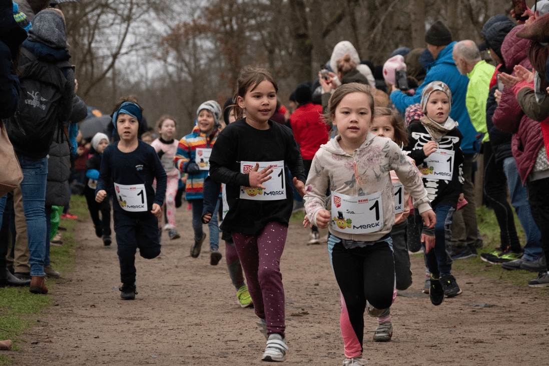Gruppe von Kindern beim Wettlauf auf einem staubigen Weg, Zuschauer applaudieren am Rand. Alle tragen Startnummern.