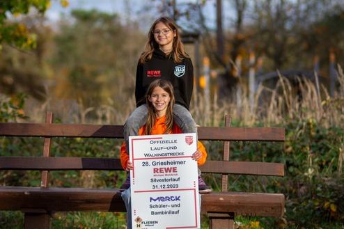 Zwei Kinder sitzen auf einer Bank im Freien, eines steht hinter dem anderen und hält ein Schild mit Laufveranstaltungsdetails.