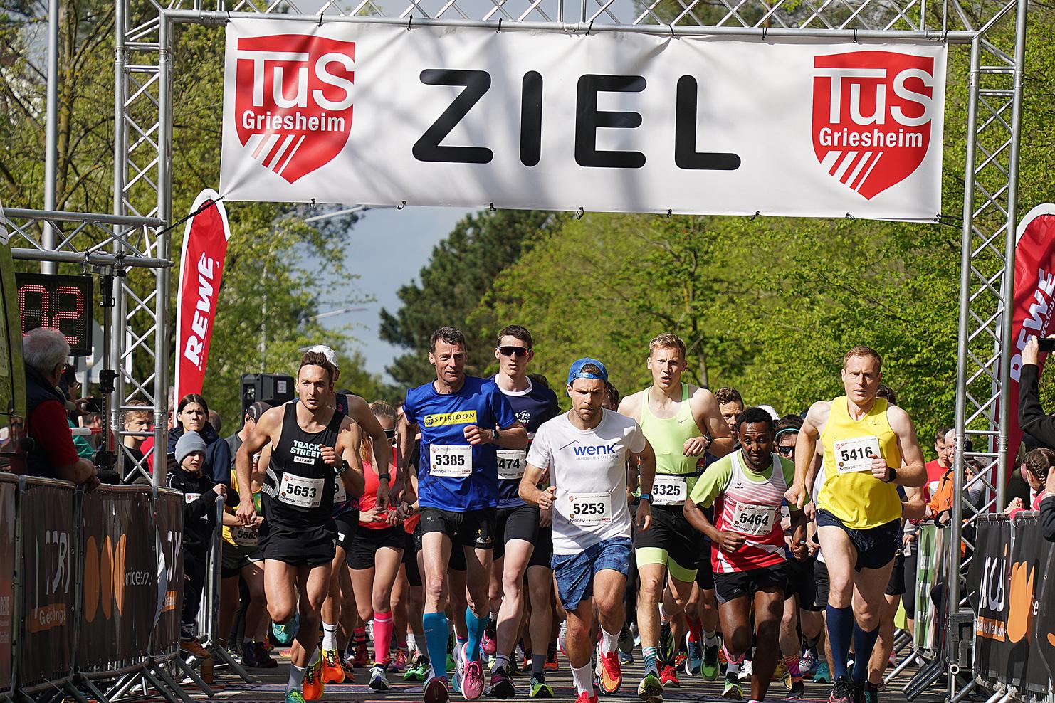 Gruppe von Läufern beim Start eines Rennens unter einem Zielbanner in Griesheim, umgeben von Zuschauern und Bäumen.