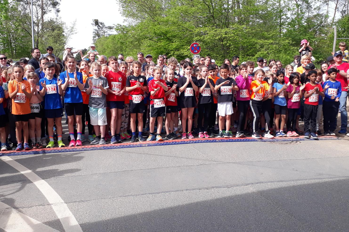 Gruppe von Kindern in Sportbekleidung mit Startnummern stehen vor einem Wettkampf, Publikum im Hintergrund.