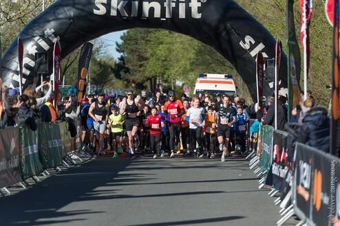 Start des Laufwettbewerbs mit zahlreichen Teilnehmern und Zuschauern unter einem großen, angedeuteten Zielbogen.