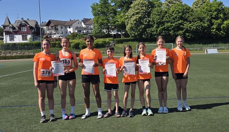Gruppe von sieben Jugendlichen in orangefarbener Sportkleidung, die Urkunden auf einem Sportplatz halten.