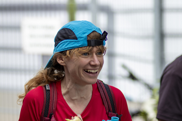 L&auml;chelnde Frau mit blauer M&uuml;tze und rotem Shirt, h&auml;lt einen Snack in der Hand, in einer lebhaften Umgebung.