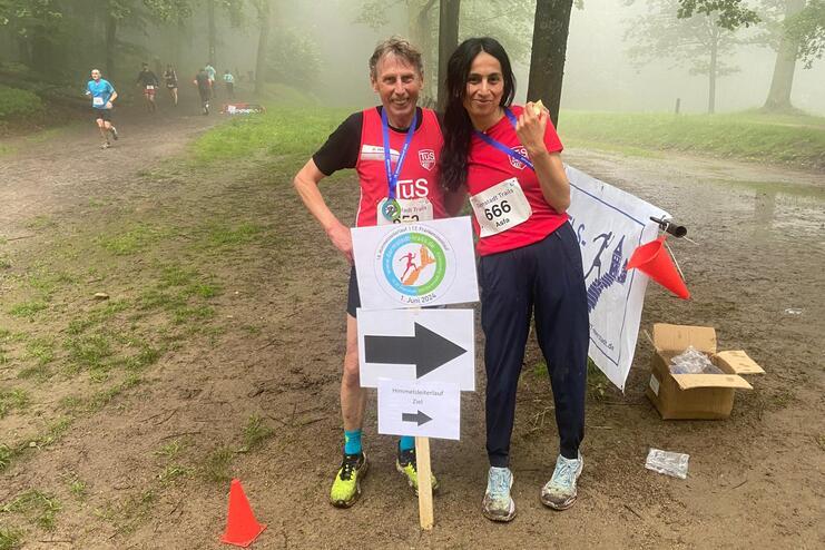 Zwei Sportler in roten Shirts posieren neben einem Wegweiser w&auml;hrend eines Laufevents in nebliger Waldlandschaft.