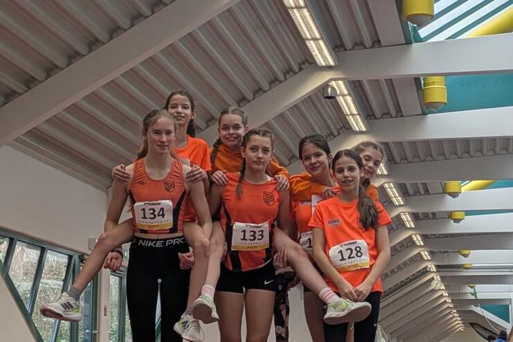 Sieben Athletinnen in orangefarbener Sportbekleidung posieren fröhlich gemeinsam auf einer Laufbahn in einer Sporthalle.