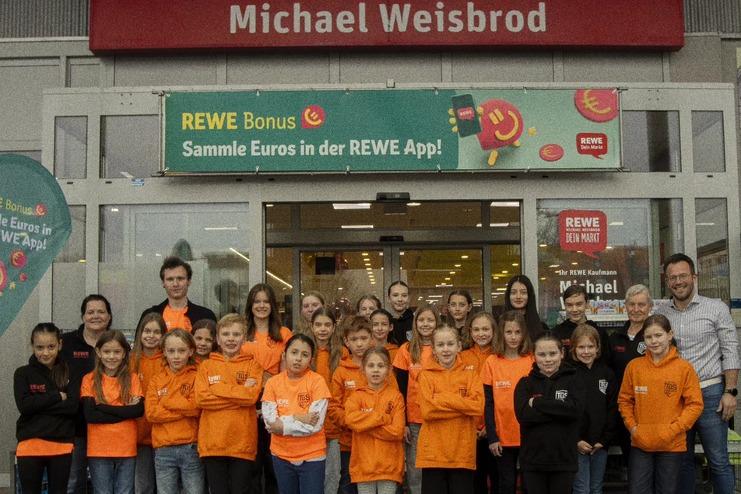 Gruppe von Kindern und Erwachsenen in orangenen und schwarzen T-Shirts vor einem REWE-Markt.