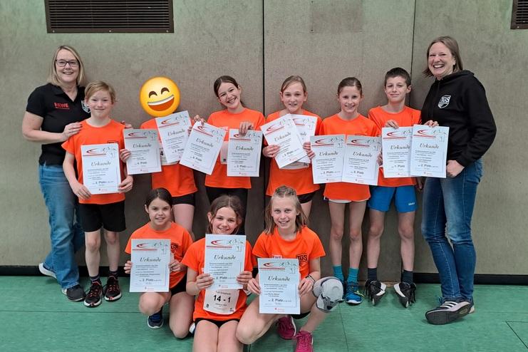 Gruppe von Kindern in orangenen T-Shirts hält Urkunden für sportliche Leistungen in einer Sporthalle.