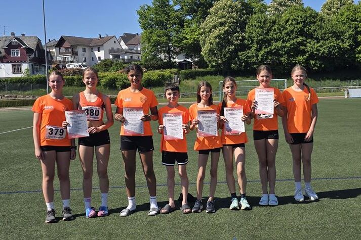 Gruppe von sieben Jugendlichen in orangefarbener Sportkleidung, die Urkunden auf einem Sportplatz halten.
