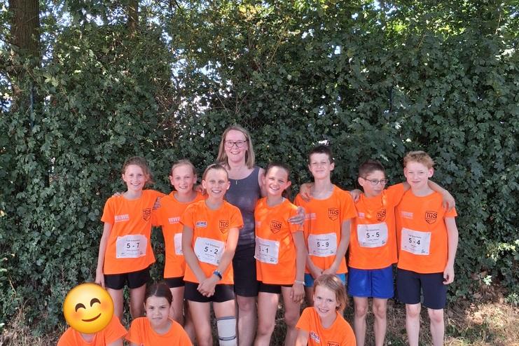 Gruppe von zehn Jungen und Mädchen in orangefarbenen T-Shirts mit Nummern, posierend vor einer grünen Hecke.