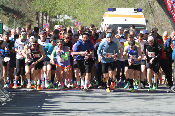 Start einer Laufveranstaltung mit zahlreichen L&auml;ufern, die sich auf den Wettkampf konzentrieren. Ein Krankenwagen im Hintergrund.