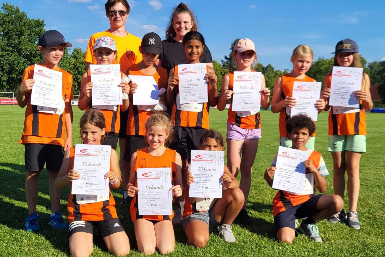 Gruppe von Kindern in Sportkleidung hält Urkunden auf einem Rasenplatz, im Hintergrund blauer Himmel und Bäume.
