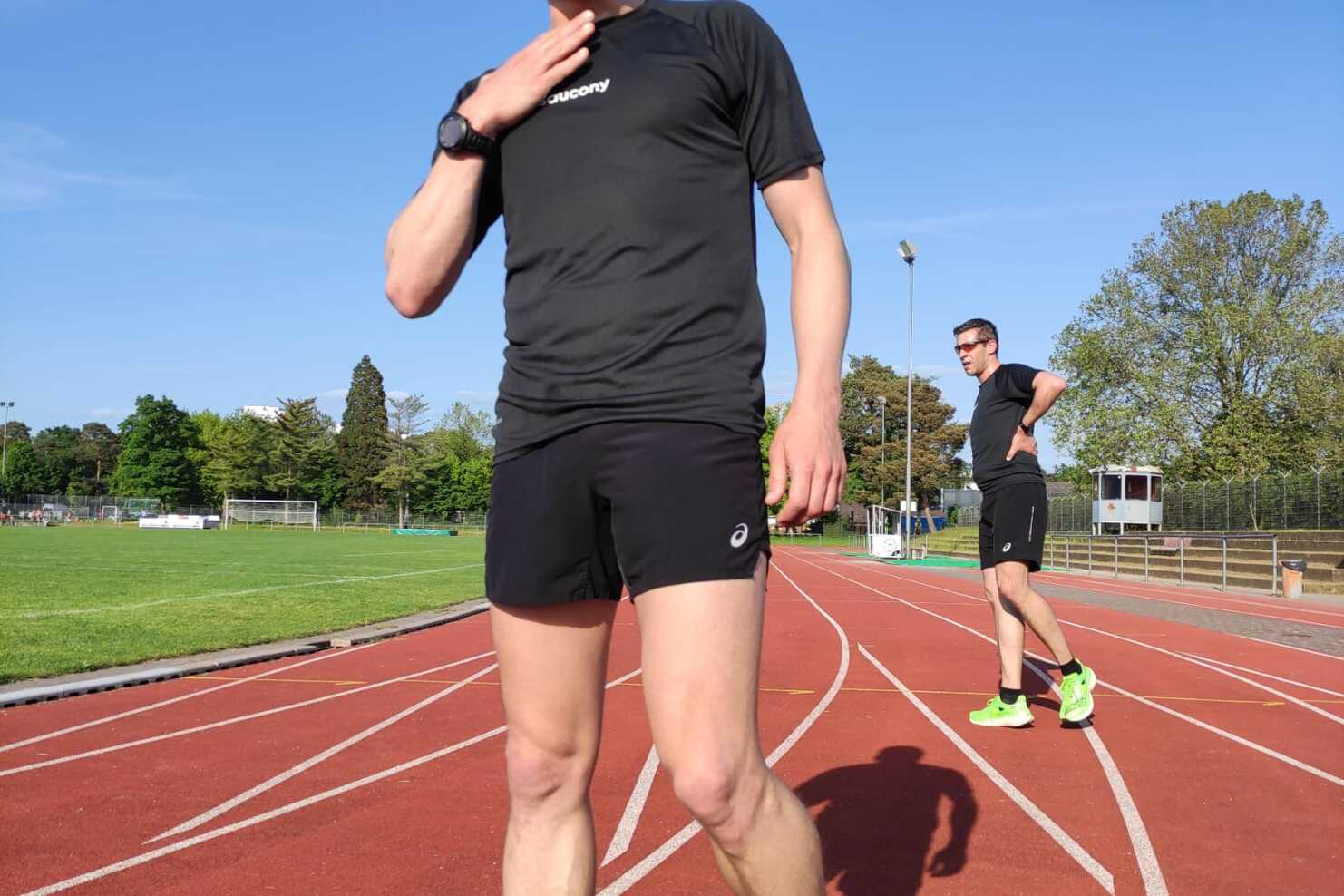 Junger Mann in Sportkleidung auf einer roten Laufbahn steht lächelnd, ein weiterer Läufer im Hintergrund. Klare Wolken am Himmel.