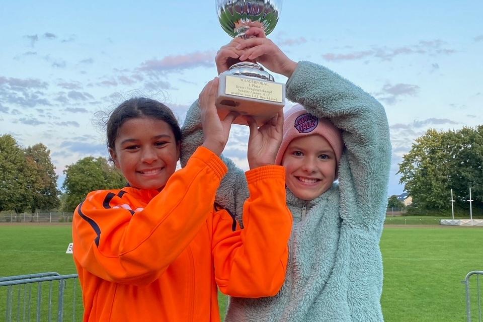 Zwei Mädchen in Sportbekleidung feiern mit einem Pokal auf einem grünen Sportfeld unter blauem Himmel.