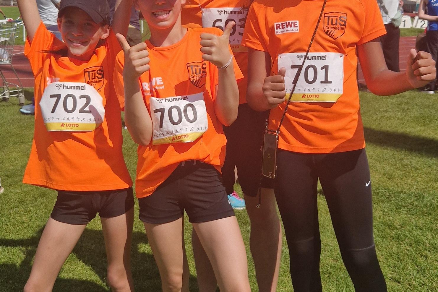 Vier Kinder und ein Jugendlicher in orangen Sporttrikots posieren fröhlich auf einem Rasenfeld nach einem Wettkampf.