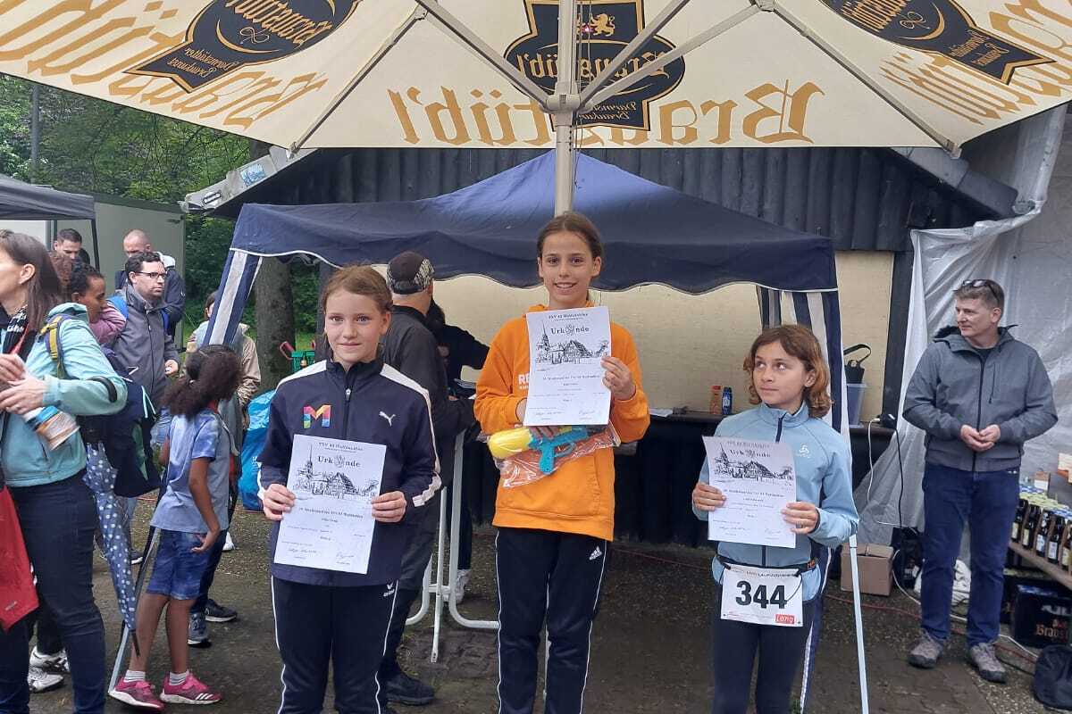 Zwei Mädchen stehen auf dem Siegertreppchen und halten Urkunden in den Händen, umgeben von Zuschauern unter einem Sonnenschirm.