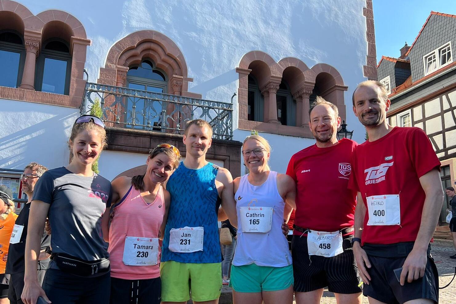 Sechsergruppe von Läufern vor einem historischen Gebäude, eines trägt ein rotes Shirt, die anderen sportliche Outfits.