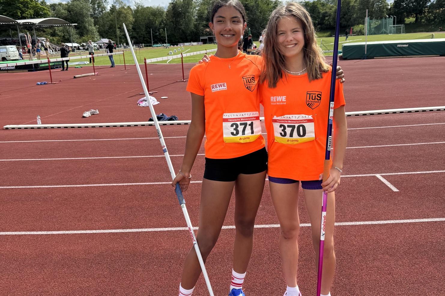 Zwei Mädchen in orangenen Trikots halten Speere auf einer roten Laufbahn unter blauem Himmel.