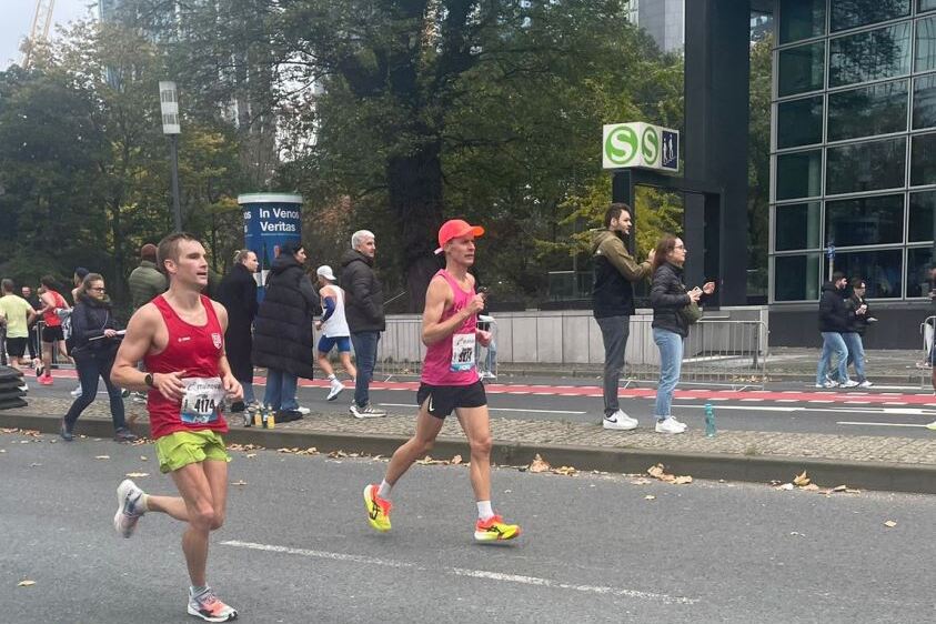 Zwei Läufer in Sportkleidung während eines Marathons, umgeben von Zuschauern und urbaner Umgebung.