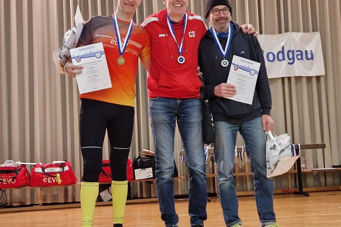 Drei Sportler stehen auf einer Bühne, tragen Medaillen und halten Urkunden in der Hand. Hintergrund mit Vorhängen.