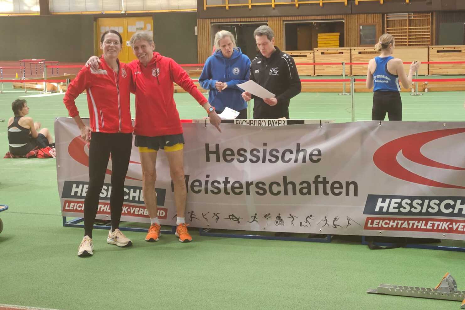 Zwei Athleten in Sportkleidung posieren lächelnd vor einem Banner der Hessischen Meisterschaften in einer Sporthalle.