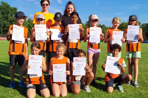 Gruppe von Kindern in Sportkleidung hält Urkunden auf einem Rasenplatz, im Hintergrund blauer Himmel und Bäume.