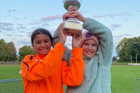 Zwei Mädchen in Sportbekleidung feiern mit einem Pokal auf einem grünen Sportfeld unter blauem Himmel.