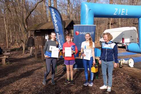 Vier Läuferinnen mit Urkunden stehen vor einem Zielbogen im Wald, während eine Person im Hintergrund sitzt.