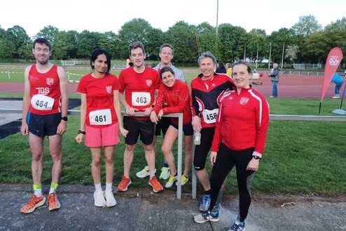 Gruppe von sieben Athleten in roten Trikots mit Wettbewerbsnummern auf einem Sportplatz, im Hintergrund eine Laufbahn.