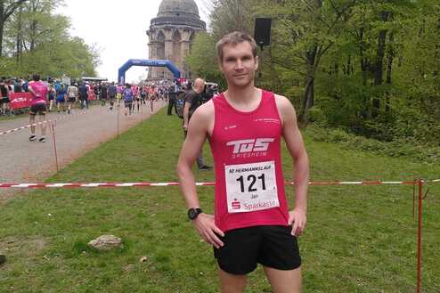 Läufer in Sportbekleidung mit Startnummer 121 posiert vor dem Hermannsdenkmal und einer Menschenmenge auf dem Weg.