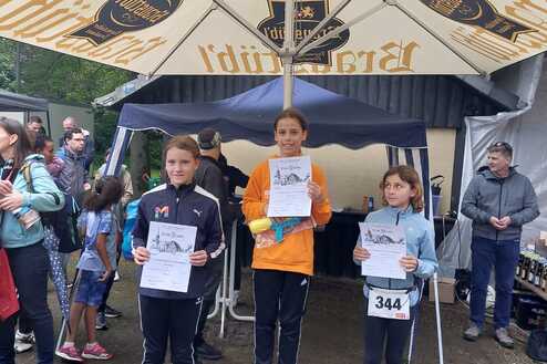 Zwei Mädchen stehen auf dem Siegertreppchen und halten Urkunden in den Händen, umgeben von Zuschauern unter einem Sonnenschirm.