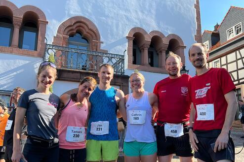 Sechsergruppe von Läufern vor einem historischen Gebäude, eines trägt ein rotes Shirt, die anderen sportliche Outfits.