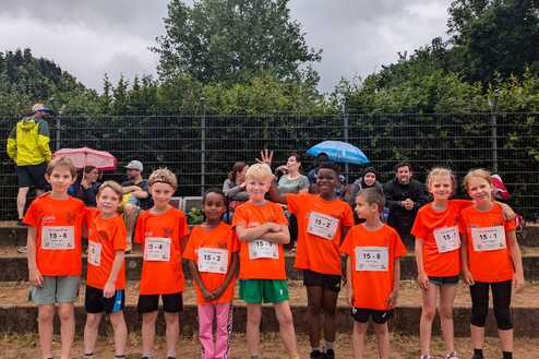 Gruppe von Kindern in orangen T-Shirts mit Startnummern, die fröhlich vor einer Zuschauerbank stehen.