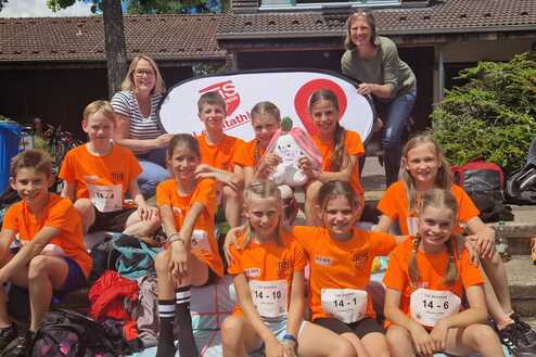 Gruppe von Kindern in orangefarbenen T-Shirts sitzt vor einem Banner, umgeben von Sportausrüstung und Rucksäcken.