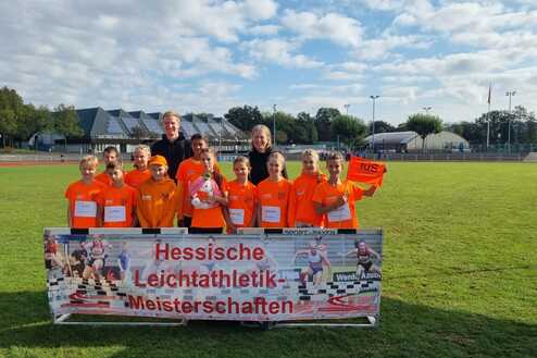 Gruppe junger Sportler in orangen Trikots posiert vor einem Banner der Hessischen Leichtathletik-Meisterschaften.