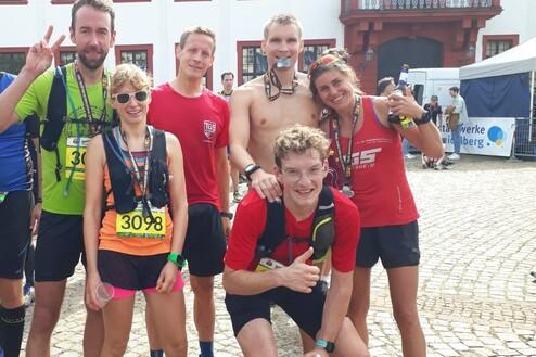 Gruppe von sechs Trailrunnern mit Medaillen lächeln vor einem Gebäude nach dem Wettkampf.