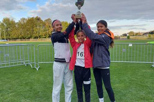 Drei Mädchen halten gemeinsam einen Pokal, stehend auf einem Sportplatz mit grüner Wiese und blauem Himmel.