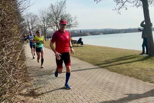 Jogger in rotem Shirt und blauen Socken läuft auf gepflastertem Weg am Wasser entlang, weitere Läufer im Hintergrund.