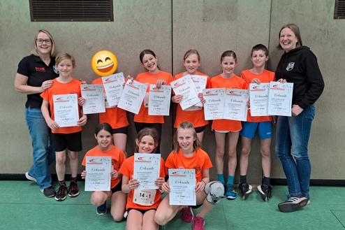 Gruppe von Kindern in orangenen T-Shirts hält Urkunden für sportliche Leistungen in einer Sporthalle.