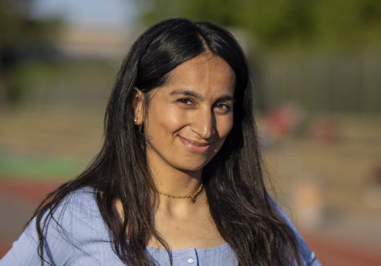Nahaufnahme einer lächelnden Frau mit langen, dunklen Haaren in einem blauen Oberteil, unscharfer Hintergrund.