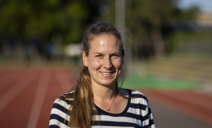 Portrait einer lächelnden Frau mit gestreiftem Shirt, Hintergrund mit einer Laufbahn und verschwommenen Sportanlagen.
