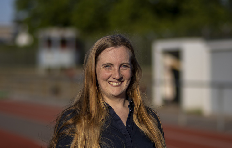 Porträt einer lächelnden Frau mit langen, blonden Haaren, im Hintergrund eine Sportanlage mit Laufbahn und Bauten.
