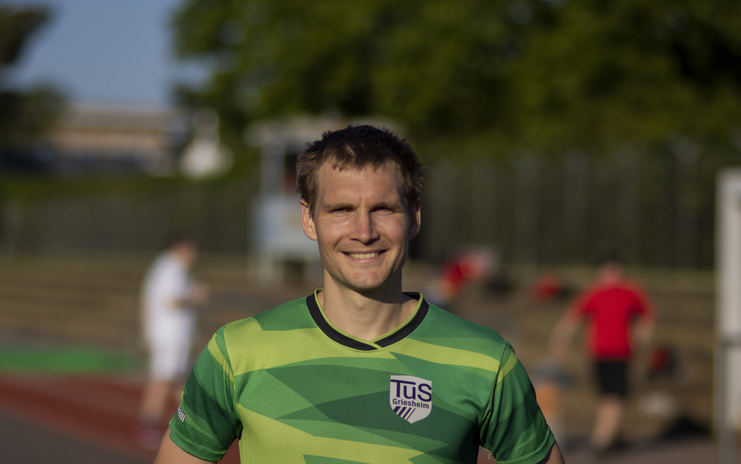 Lächelnder Mann in grünem Trikot von TuS Griesheim steht im Vordergrund eines Sportplatzes, im Hintergrund andere Sportler.
