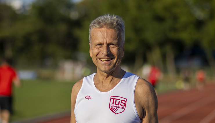 Lächelnder männlicher Läufer in Trainingstrikot mit Vereinslogo auf einer Laufbahn im Hintergrund.