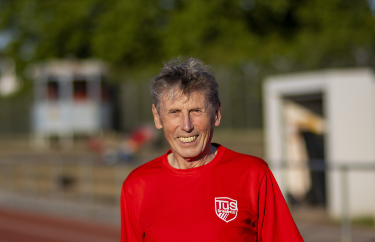 Lächelnder älterer Mann in rotem Sporttrikot steht auf einem Sportplatz mit unscharfen Hintergrund.
