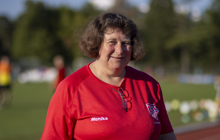 Frau mit lockigem, braunem Haar trägt ein rotes T-Shirt und lächelt auf einem Sportplatz im Hintergrund.