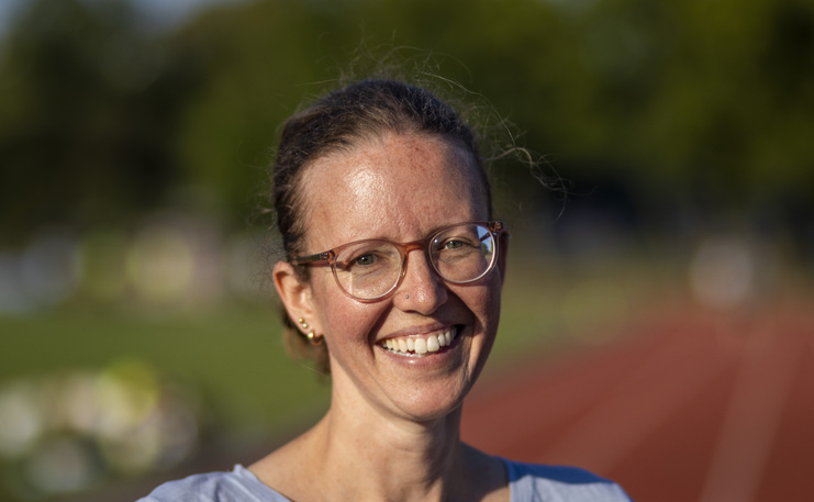 Lächelnde Frau mit Brille, stehend vor einer Laufbahn in einem Stadion. Hintergrund unscharf mit grünem Rasen.